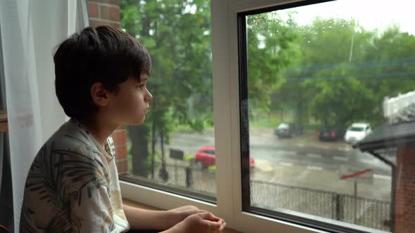 A boy sits at a wet window during the rain and looks at the street