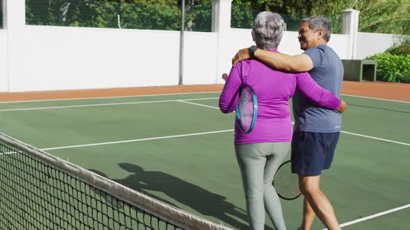 Video of happy biracial senior couple embracing and walking with rackets on tennis court