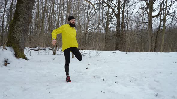 Bearded Fit Sports Man in Yellow Coat Stretching Legs before Running in Winter Forest