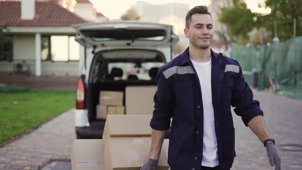 Young Handsome Smiling Caucasian Delivery Man with Boxes on Cart Going From Van with Opened Truck