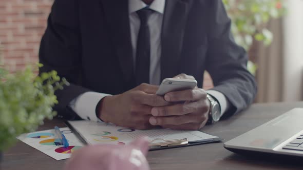 Black Man in an Official Suit Prints at Phone at an Office Table with a Laptop and Documents Closeup