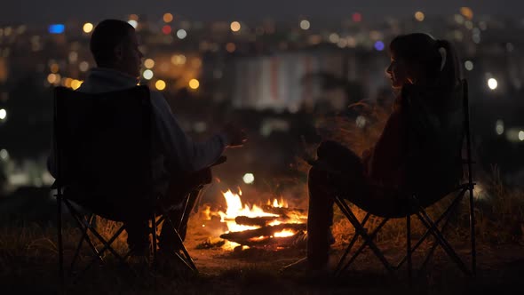 Young couple in love talking with each other sitting on chairs near bonfire at night enjoying 