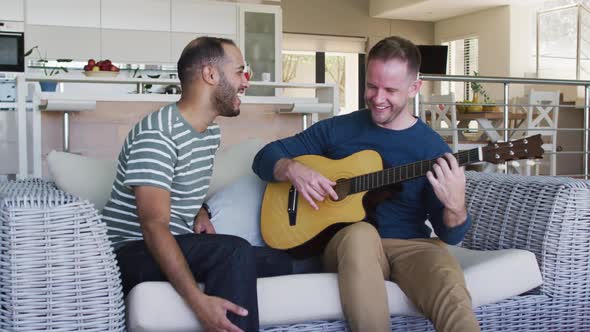 Multi ethnic gay sex couple sitting on couch one playing guitar