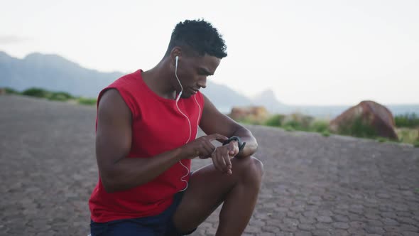 African american man wearing earphones using fitness band while sitting on the road