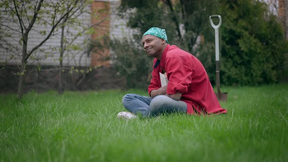 Side View Wide Shot of Happy Relaxed African American Man Sitting on Green Grass in Summer Garden