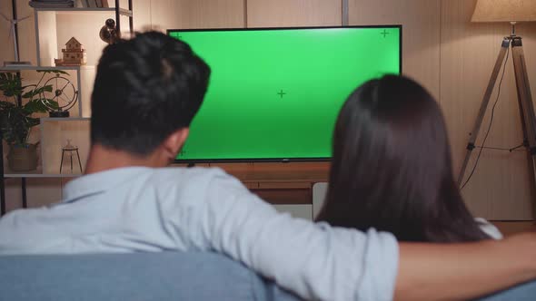 Back View Of Young Asian Couple Relaxing And Watching Tv With Mock Up Green Screen At Home