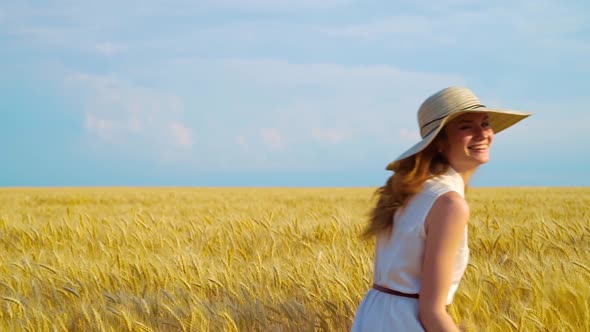 Happy Girl in Light Dress and Straw Hat Walking on Wheat Field in Harvest Season