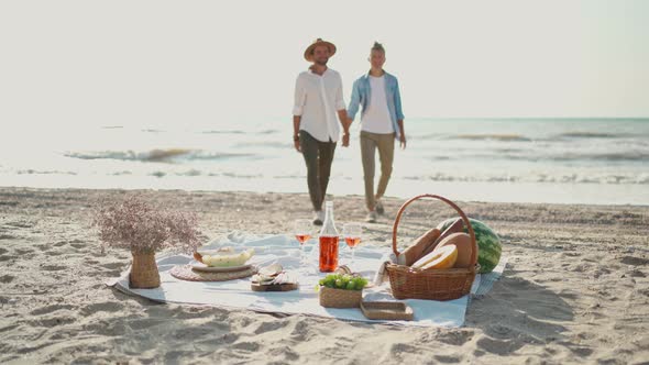 Silhouette Gay Couple Walking By Sea Beach Focus on Picnic Blanket with Wine Glasses and Food