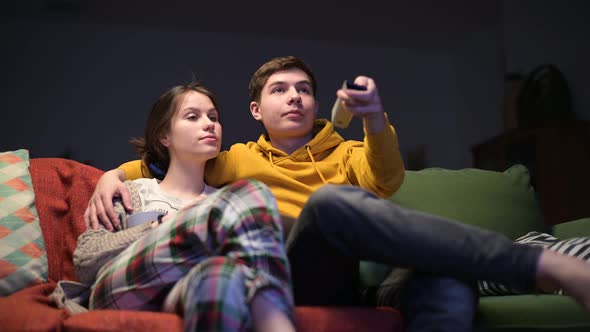 A young guy and his girlfriend sit on the sofa and turn on the TV in the evening