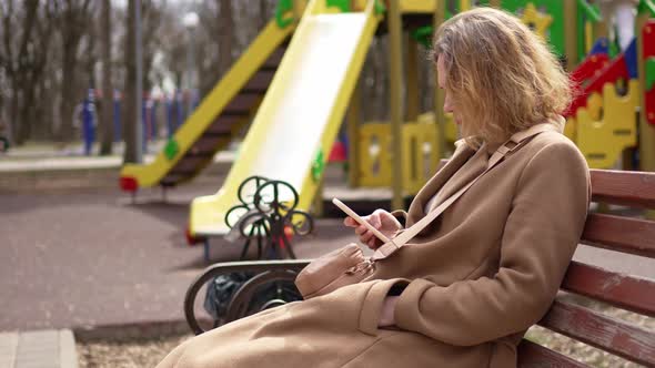 a Woman in a Brown Coat Sit on a Bench with Smartphones