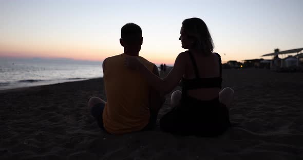 Husband and Wife are Sitting on Beach Daughter Runs Up to and Hugs Them at Sunset