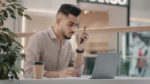 Serious Focused Arab Guy Sitting at Table Typing on Modern Laptop Answering Phone Call Communicate