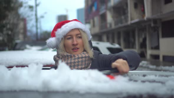 young beautiful Caucasian woman in Santa hat cleans car from snow with a brush