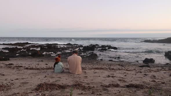 A father and daughter sit on a cold sandy southern Australian beach together and watch the sunset.