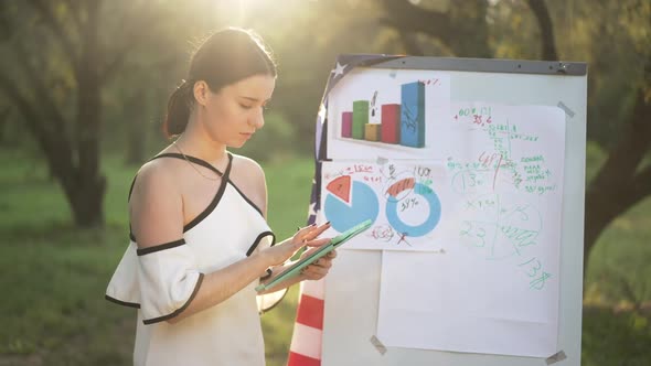 Young Thoughtful Woman Surfing Internet on Tablet Analyzing Graphs in Sunshine Standing in Park