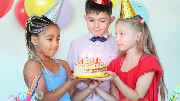 Little Girls Hold Cake While Boy Blows Candles at Birthday