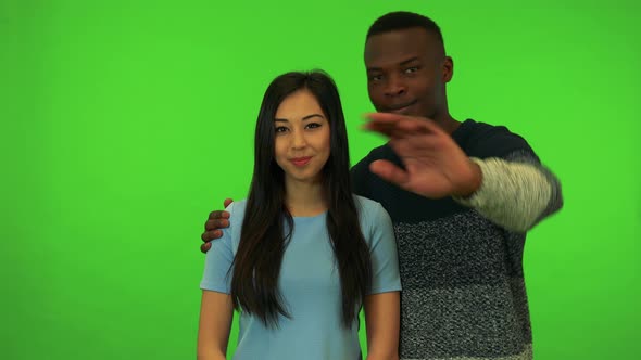 A Young Asian Woman and a Young Black Man Wave at the Camera with a Smile - Green Screen Studio