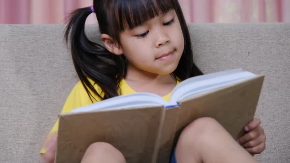 Cute little girl reading a book while sitting on the sofa at home.