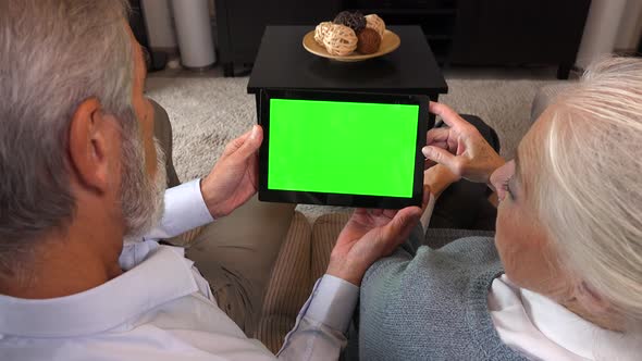 An Elderly Couple Sits on A Couch in An Apartment and Looks at A Tablet with A Green Screen