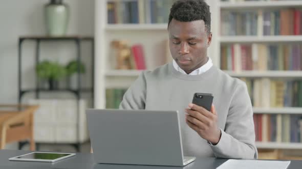 African Man Using Smartphone While Using Laptop in Library