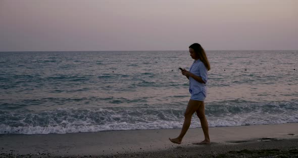 Lonely Brunette Woman Is Walking on Shore of Sea After Sunset, Looking on Screen of Mobile Phone