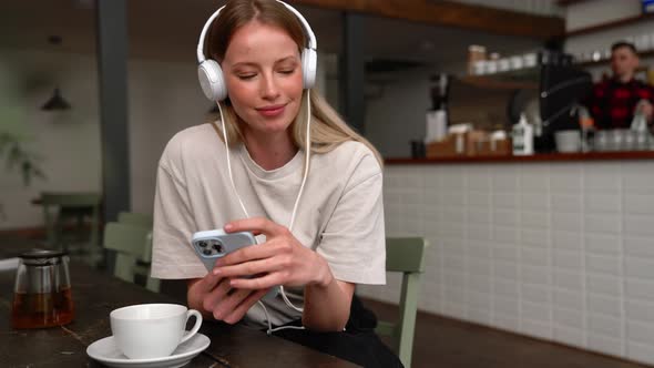 Positive blonde woman listening music in headphones and typing by phone