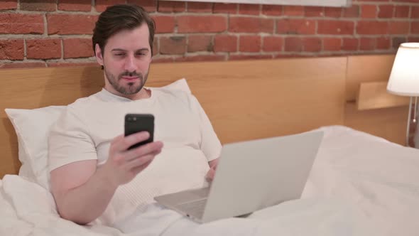 Casual Young Man with Laptop Using Smartphone in Bed