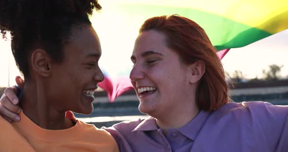Multiracial female couple kissing together at sunset while holding LGBT rainbow flag