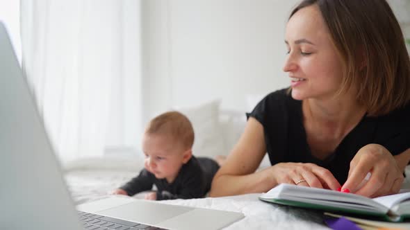 Young Mother Freelancer with Baby Son Sitting on Bed and Trying to Work at Laptop