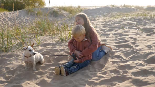 Brother and Sister in a Hoodie Sit Their Dog on the Sand