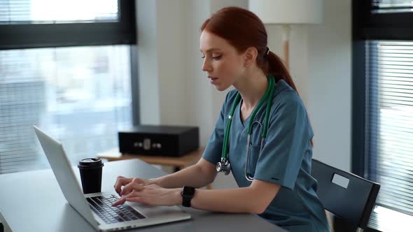 Attractive Young Female Practitioner Doctor in Blue Green Medical Uniform Working Typing on Laptop
