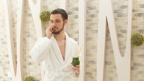 Attractive Man in White Robe Spends Time in Elite Spa Center. Talking on Mobile Phone and Walks Past