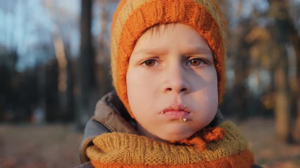 Boy in a Knitted Hat and a Snood Chewing Food with His Mouth Closed in an Autumn Park