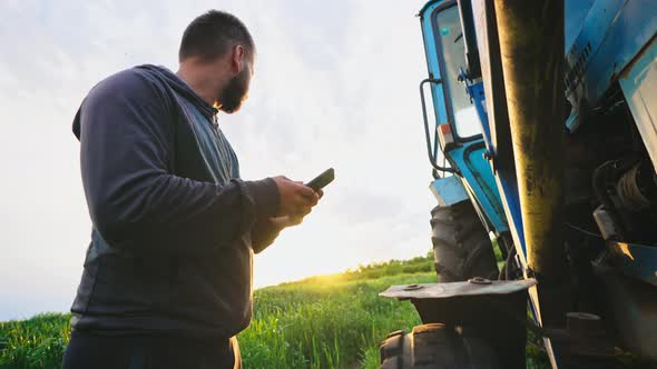 Man in Field with Phone in His Hands Stands By His Tractor and Knocks on Wheel Checking Condition of