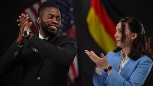 Clapping Caucasian Woman Inviting African American Man to the Stage in Camera Flashes at Press