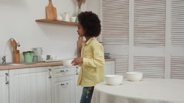 Little African American Girl Removes Deep White Bowls From Table and Places Them in Sink to Wash