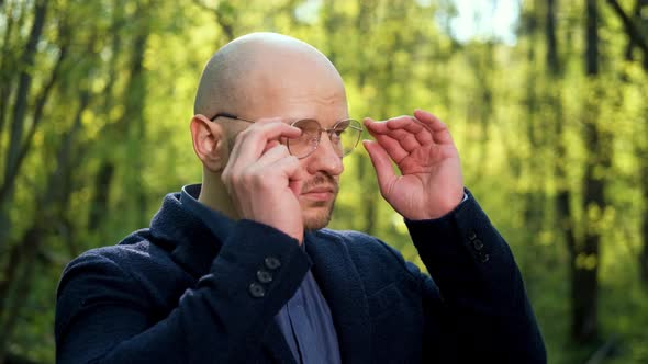 A Bald Man Puts on Glasses Turns His Head and Looks at the Camera in the Forest