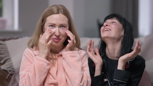 Two Joyful Beautiful Women Applying Eye Patches Laughing Talking in Slow Motion Sitting at Home