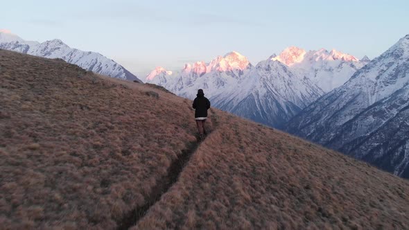 An Aerial View of a Young Girl in a Cap Runs Down a Mountain Hill Towards the Snow-capped Mountains