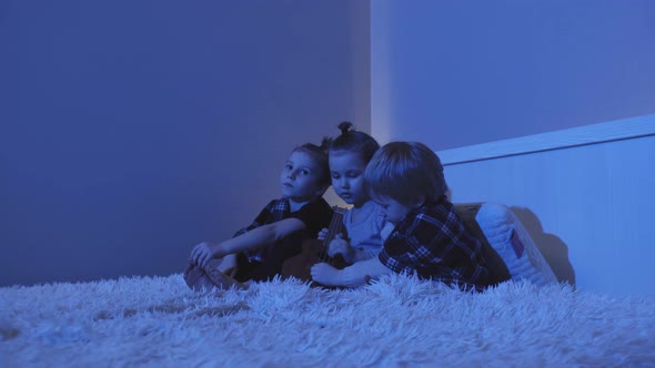 Three Little Boys Sit on Bed and Play Guitar in Room at Night