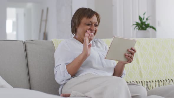 Happy african american senior woman sitting on couch making video call using tablet, waving