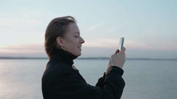 A Woman Takes Pictures on Her Phone of a Lake at Sunset on an Autumn Evening