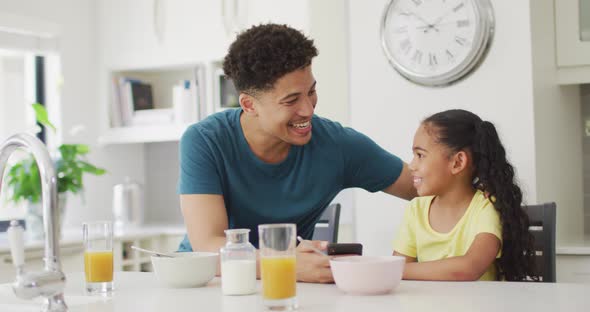 Happy biracial father and daughter eating breakfast and using smartphone together