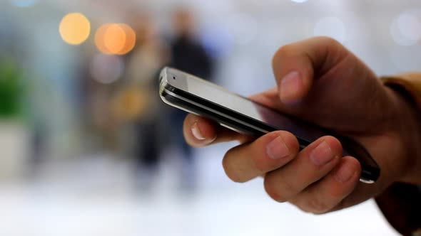 A Man's Hand Uses Black Smartphone on the Background Walking People and Bokeh Lights