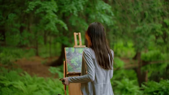 Rear View of a Young Artist Female Painting a Landscape and Wipes the Brush in a Summer Forest