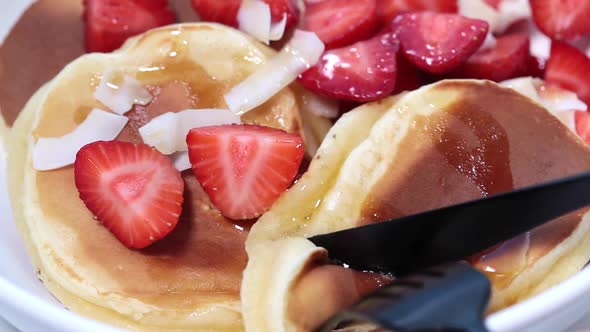 Homemade pancakes with strawberries and coconut in white bowl, cut with a knife and fork.