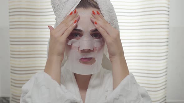 Cheerful Young Woman looking in the Camera Applying the Sheet Mask on Her Face in the Bathroom