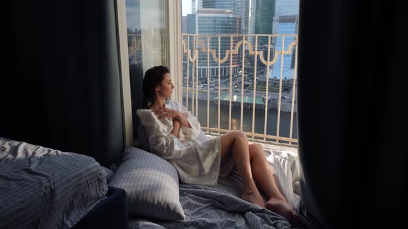 Woman in a Bathrobe Sits on the Floor of a Balcony Against the Backdrop of a Skyscraper During the