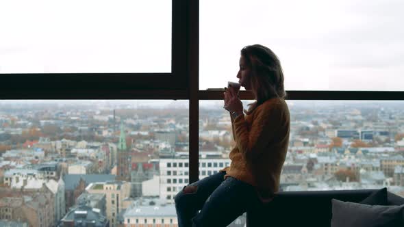 Young Woman Is Sitting Against a City View and Drinking Coffee