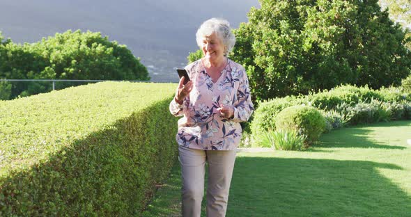 Caucasian senior woman smiling while talking on smartphone while walking in the garden